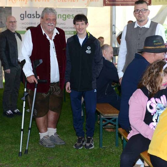 Zwei Männer, einer mit einer Krücke und der andere mit einer schwarzen Jacke mit Logo, stehen auf einem Rasen. Andere Leute sitzen auf Bänken.