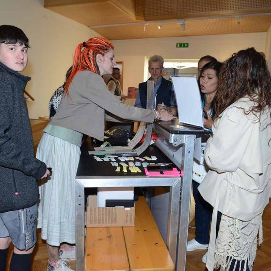 Eine Gruppe von Menschen befindet sich in einem Raum mit einem Tisch, und eine Frau in einem weißen Kleid steht hinter dem Tisch und hält ein weißes Objekt. Ein Mann und eine Frau stehen vor dem Tisch. Eine Frau mit roten Haaren schaut sich das Objekt an. Ein Mann steht hinter der Frau. Der Raum hat eine Holzwand und eine weiße Decke.