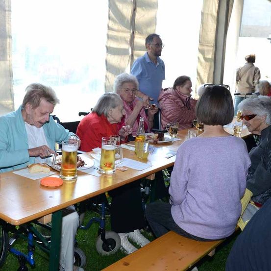 Ältere Menschen sitzen an einem Tisch, essen, trinken Bier und lachen. Ein Mann steht im Hintergrund hinter ihnen.