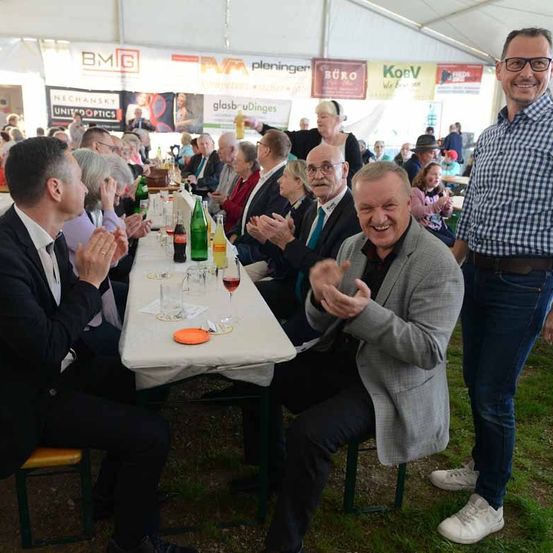 Eine Gruppe von Menschen sitzt an einem langen Tisch, vielleicht in einem Zelt, und sie klatschen in die Hände.