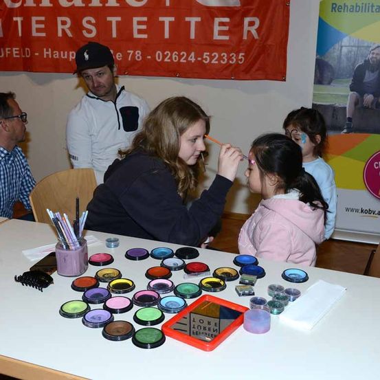 Eine Frau malt das Gesicht eines jungen Mädchens mit Gesichtsfarbe bei einer Veranstaltung. In der Nähe sind andere Leute, und ein Tisch mit verschiedenen Farben von Gesichtsfarbe.