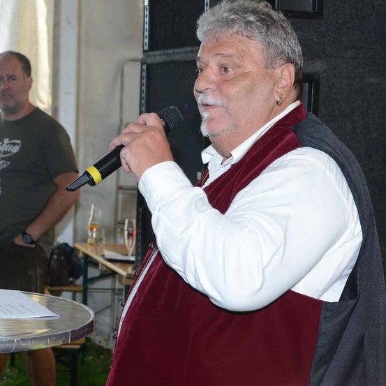 Ein Mann spricht in ein Mikrofon bei einer Veranstaltung im Freien. Ein Mann in einem grünen T-Shirt steht hinter ihm.