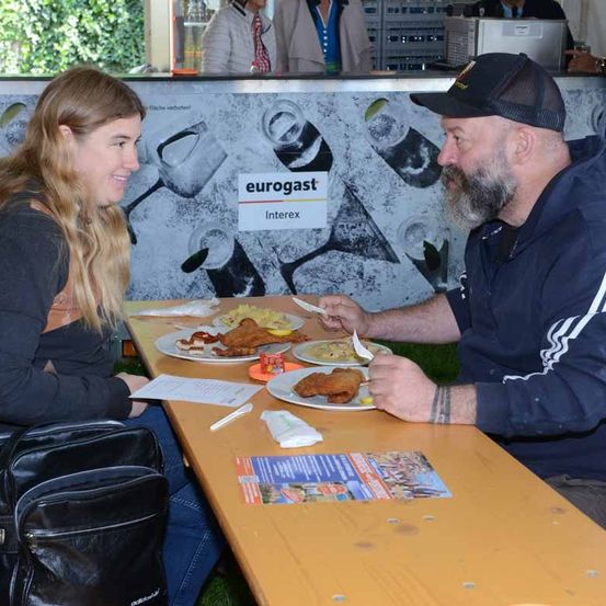 Ein Mann und eine Frau sitzen an einem Tisch und essen. Der Mann trägt eine Baseballkappe und hat einen Bart. Die Frau trägt ein schwarzes Oberteil.
