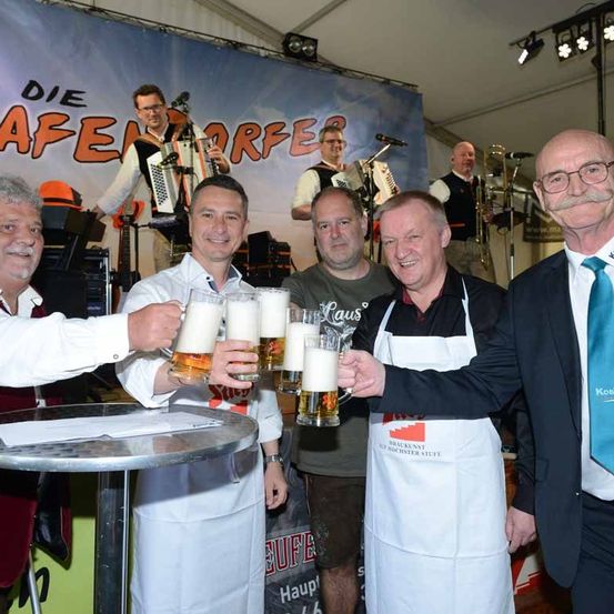 Fünf Männer in weißen Schürzen halten Gläser mit Bier vor einem Banner mit der Aufschrift 'Die Frauendorfer'. Dahinter spielen Musiker Instrumente, darunter Akkordeon und Posaune.