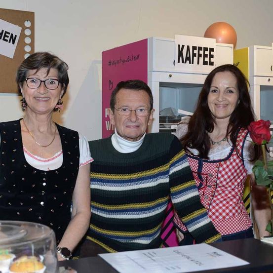 Drei Personen stehen hinter einem Tisch mit Cupcakes in einer Vitrine. Die Frau links trägt eine Brille und eine Halskette. Der Mann in der Mitte trägt ein Rollkragenshirt und einen gestreiften Pullover. Die Frau rechts trägt eine rote und weiße Schürze und hält eine Rose.