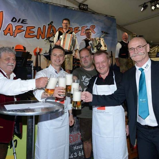 Fünf Männer in Schürzen stoßen mit Biergläsern an. Zwei Musiker spielen Akkordeons hinter ihnen. Ein Banner mit der Aufschrift 'Die Abendoper'. Ein Schild auf dem Tisch mit der Aufschrift 'Haup 43 66'.