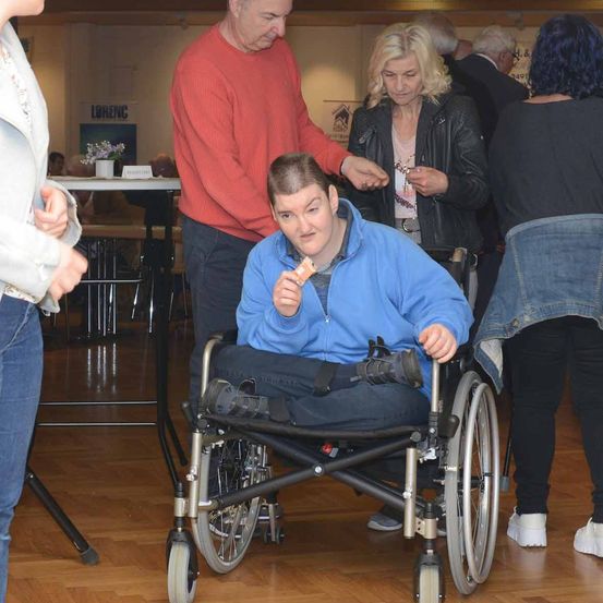 Eine junge Frau im Rollstuhl isst einen Snack, während ein Mann und eine Frau hinter ihr zu sprechen scheinen. Die Szene spielt drinnen mit Holzböden und mehreren Personen im Hintergrund.