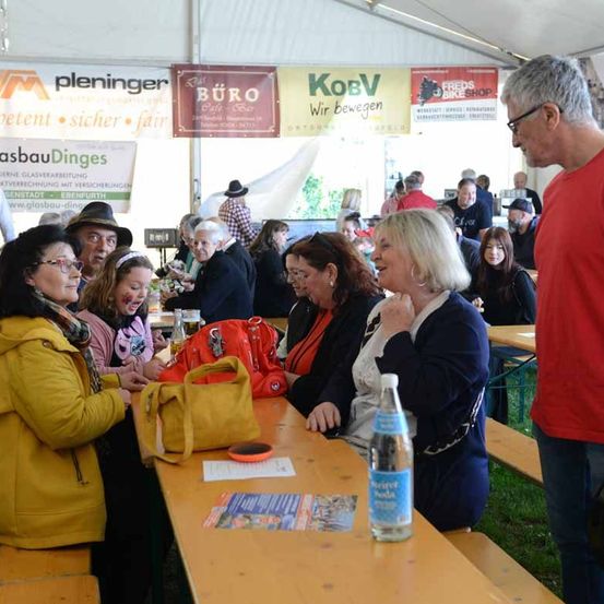 Eine Gruppe von Menschen sitzt um einen Tisch in einem Zelt. Der Tisch hat eine Flasche, eine rote Tasche und einige Papiere. Ein Mann in einem roten T-Shirt steht neben ihnen.