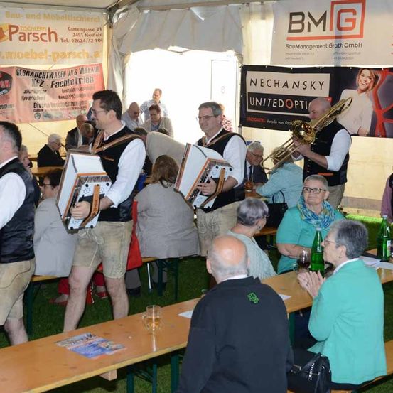 Eine Gruppe von Musikern tritt bei einer Veranstaltung im Freien auf. Die Musiker spielen Akkordeons und eine Posaune. Die Menschen sitzen an Tischen und genießen die Aufführung. Im Hintergrund sind Banner mit Werbung angebracht.