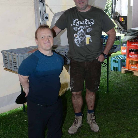 Zwei Männer stehen auf einem Grasfeld, wahrscheinlich bei einer Veranstaltung im Freien. Der Mann auf der linken Seite trägt ein blaues Shirt und schwarze Hose. Der Mann auf der rechten Seite trägt ein grünes Shirt mit Kuh- und Bierglas-Design und braune Shorts. Hinter ihnen befinden sich Kisten und ein Tisch mit einem schwarzen Hut.