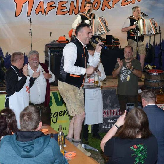 Eine Gruppe von Menschen steht auf einer Bühne und tritt auf. Ein Mann hält ein Mikrofon und scheint zu singen. Hinter ihm spielen einige Leute Musikinstrumente. Ein Mann, der neben dem Mikrofon steht, scheint zu klatschen. Ein paar Leute stehen vor der Bühne und ein Fass ist dahinter aufgestellt.