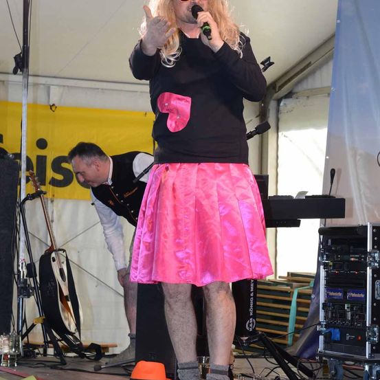 Ein Mann auf der Bühne in einem schwarzen Shirt mit einer pinken Nummer 7 und einem pinken Rock, singt in ein Mikrofon.