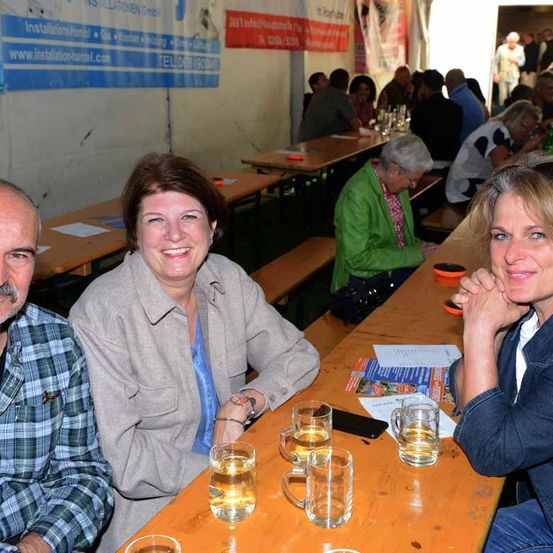 Mehrere Personen versammeln sich an einem Tisch in einem Restaurant, lächeln und posieren für ein Foto. Hinter ihnen sind Tische mit sitzenden und stehenden Personen zu sehen. An der Wand befinden sich mehrere Schilder, eines davon trägt den Schriftzug 'Installations-Handel'.
