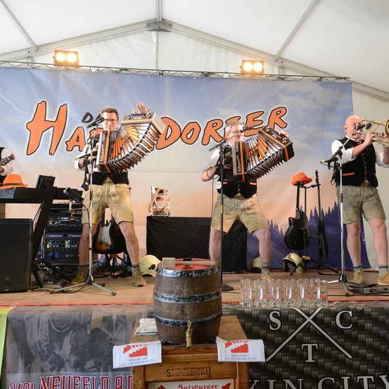 Drei Männer spielen auf der Bühne mit Musikinstrumenten, darunter Trompete, Akkordeon und Trommeln. Auf der Bühne stehen ein Fass und Gläser.