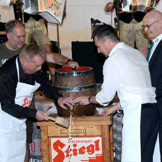 Mehrere Männer stehen um einen Holzfass herum. Ein Mann klopft mit einem Hammer auf das Fass, während ein anderer Mann einen Zapfhahn hält. Hinter ihnen spielen Musiker Akkordeon und andere Instrumente.