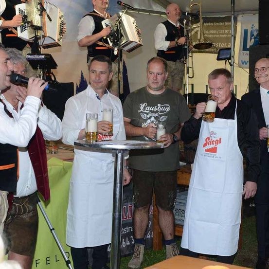 Vier Männer stehen hinter einem Tisch mit Bier. Zwei von ihnen tragen Schürzen und lächeln. Der Mann auf der linken Seite spricht in ein Mikrofon. Dahinter stehen zwei Leute, die Akkordeons spielen, ein Mann spielt Posaune und ein Mann im weißen Hemd.