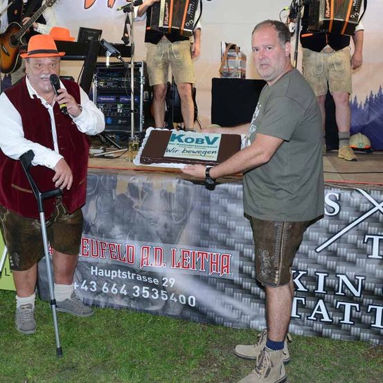 Ein Mann mit einem Gehstock hält ein Mikrofon, während ein anderer Mann einen Kuchen in der Hand hat. Dahinter stehen drei Personen auf der Bühne und spielen Musikinstrumente.
