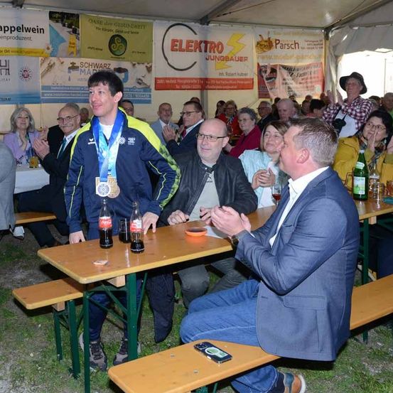 Mehrere Menschen, die um einen Tisch herum sitzen, klatschen für einen Mann in einer Medaillenjacke, der vor ihnen steht.