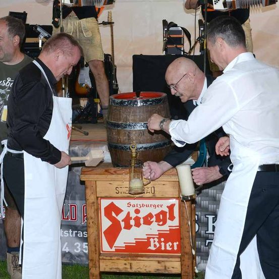 Vier Männer schenken Bier aus einem Holzfass in Gläser. Das Fass ist mit 'Stiegl Bier' beschriftet. Sie tragen Schürzen und einer trägt eine Uhr. Dahinter spielt ein Mann Gitarre.