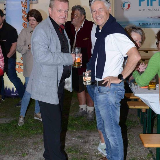 Zwei Männer stehen draußen mit Getränken in den Händen. Sie sind von anderen Personen umgeben, die um einen Picknicktisch herum sitzen und stehen. Ein Mann trägt eine Krawatte, während der andere einen Gürtel um die Taille hat.