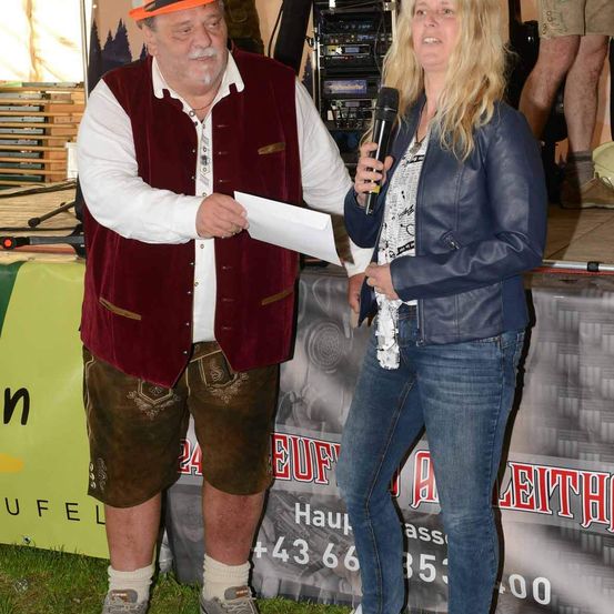 Ein Mann in einer Weste und traditionellen deutschen Lederhosen steht neben einer Frau mit einem Mikrofon. Beide lächeln und halten ein Stück Papier. Der Mann trägt einen Hut, und die Frau trägt eine Lederjacke. Hinter ihnen steht eine Bühne mit einem Hintergrund, auf dem das Wort 'Auf' in Rot steht.