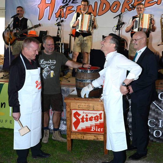 Eine Gruppe von Männern steht um einen Holzfass. Ein Mann hält einen Hammer und ein anderer trägt eine weiße Schürze. Im Hintergrund befindet sich eine Bühne mit einem Banner, Musikern und einem Publikum.