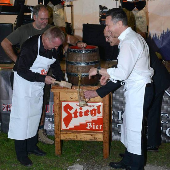 Zwei Männer in Schürzen klopfen auf ein Bierfass, um es zu öffnen, während andere zusehen.