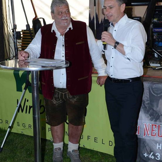 Zwei Männer stehen nebeneinander in der Nähe eines Tisches mit einem grünen Banner. Ein Mann hält ein Mikrofon und spricht, während der andere ihn ansieht.