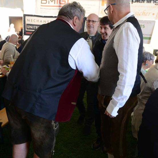 Eine Gruppe von Männern in traditioneller bayerischer Kleidung unterhält sich auf einem Rasenfeld. Der Mann auf der linken Seite trägt eine blaue Weste mit rotem Besatz, während der Mann auf der rechten Seite eine Weste über einem weißen Hemd trägt. Im Hintergrund sind weitere Personen zu sehen, und verschiedene Schilder sind links angebracht.