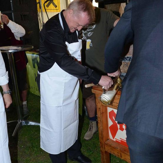Ein Mann in weißer Schürze und schwarzem Hemd schenkt ein Getränk in einen Krug bei einer Outdoor-Veranstaltung.
