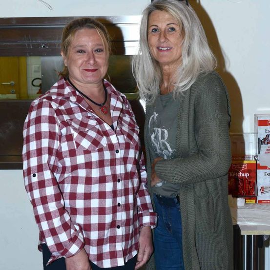 Zwei Frauen stehen lächelnd eng beieinander, eine trägt ein rot-weiß kariertes Hemd und die andere ein grünes Cape. Hinter ihnen ist ein verschwommener Hintergrund mit einem Tisch, auf dem Ketchup-Kisten stehen.