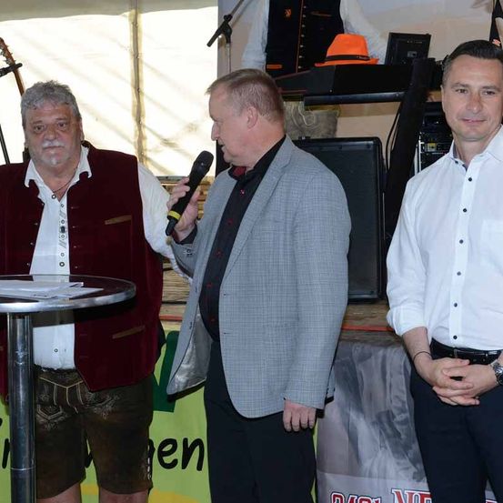 Drei Männer stehen nebeneinander vor einem Hintergrund mit dem Wort 'Den'. Der Mann auf der linken Seite trägt eine Weste, der Mann in der Mitte hält ein Mikrofon und der Mann auf der rechten Seite trägt ein weißes Hemd und eine Uhr.