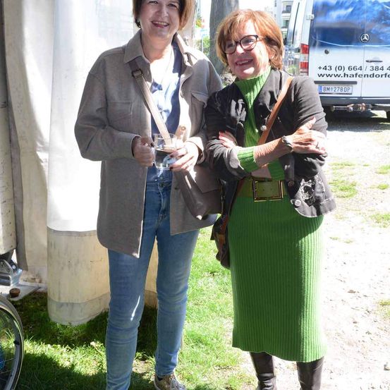 Zwei Frauen stehen im Freien, eine mit einem Getränk, lächeln. Eine trägt ein grünes Rollkragenshirt, einen Gürtel und eine Brille. Die andere trägt eine graue Jacke, Jeans und eine Uhr. Hinter ihnen steht ein weißes Zelt und ein Van.