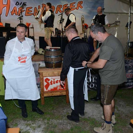 Ein Mann in einer weißen Schürze steht auf der Bühne mit einem Holzfass vor ihm. Zwei Männer richten die Schürze des Mannes in der Mitte. Ein Schild mit dem Wort 'Stiegl' steht auf dem Tisch. Dahinter spielen Musiker auf der Bühne.