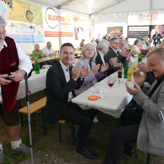 Eine Gruppe von Menschen sitzt in einem Zelt an Tischen und klatscht in die Hände. Auf den Tischen stehen Getränkeflaschen, Weingläser und Teller.