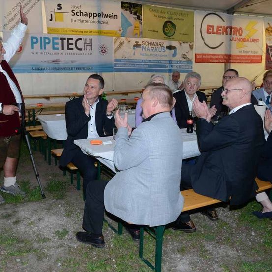 Eine Gruppe von Menschen sitzt unter einem Zelt mit verschiedenen Bannern, darunter eines für 'PIPETECH'. Sie klatschen und lächeln, während ein Mann in der Nähe ein Mikrofon hält.