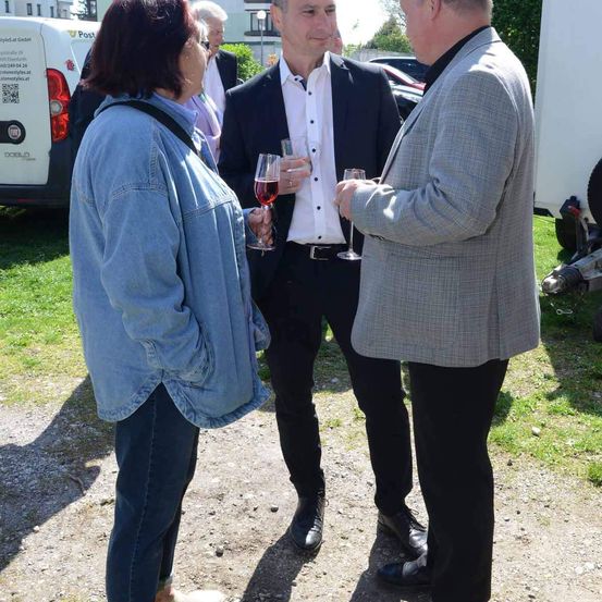 Drei Personen stehen an einem sonnigen Tag im Freien. Der Mann in der Mitte hält ein Glas Wein. Die Frau links hält ein Weinglas und trägt eine Jeansjacke. Der Mann rechts trägt einen grauen Anzug.