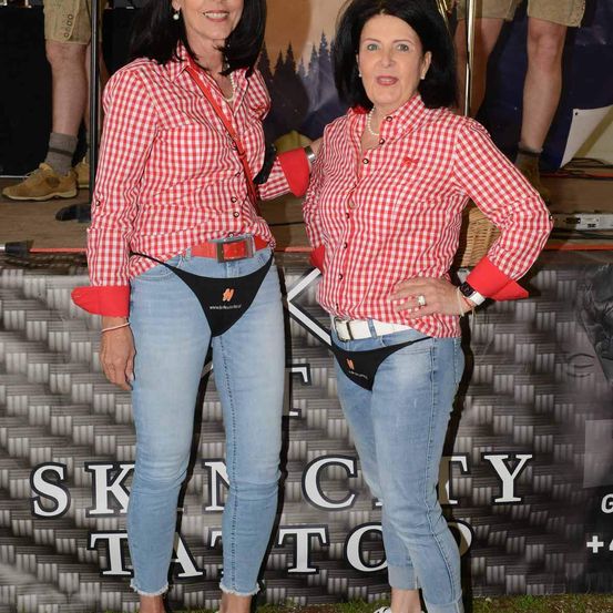 Zwei Frauen in roten und weißen karierten Hemden und Jeans stehen vor einem Schild, das Skin City Tattoo besagt. Die Frau auf der linken Seite hat einen roten Gürtel um die Taille, während die Frau auf der rechten Seite einen weißen Gürtel hat. Sie lächeln.