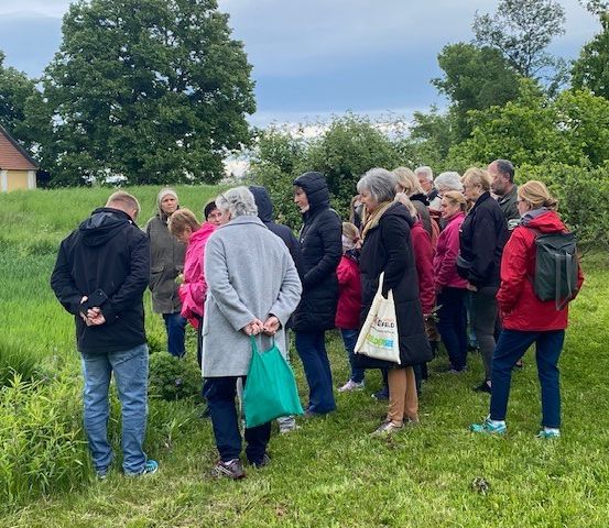 Eine Gruppe von Menschen in Jacken und Sneakern steht auf einer Rasenfläche mit Bäumen und einem Haus im Hintergrund.