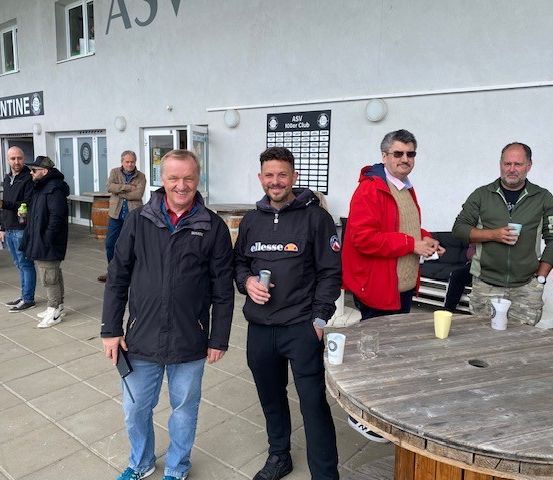 Mehrere Männer stehen vor einem Gebäude mit dem Schild 'Kantine'. Sie lächeln und halten Tassen. Der Mann in der Mitte trägt eine Jacke mit dem Wort 'ellese'.