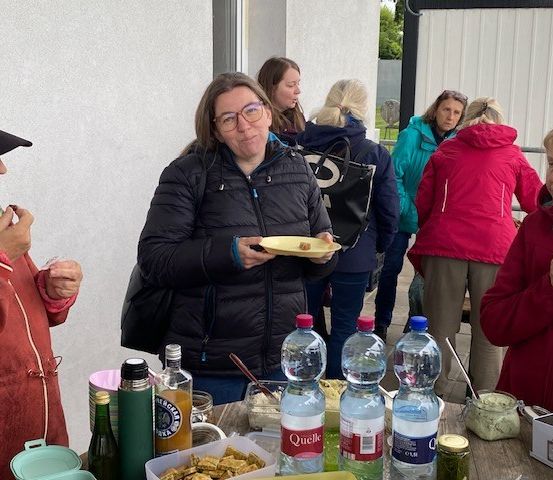 Eine Gruppe von Frauen in Jacken steht um einen Tisch mit Essen und Getränken. Eine Frau hält einen Teller mit Essen und trinkt aus einer Flasche. Der Tisch ist mit verschiedenen Flaschen, einer Box und einem Teller gefüllt. Sie befinden sich wahrscheinlich bei einer Veranstaltung im Freien.
