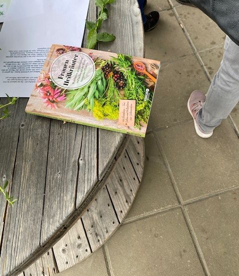Ein Buch über das Sammeln von Wildpflanzen liegt auf einem Holztisch mit einem weißen Papier darauf. Eine Person steht in der Nähe, trägt schwarze Schuhe und eine graue Tasche.