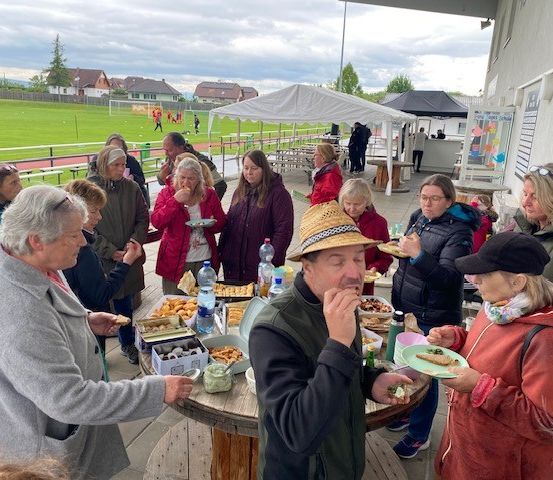 Eine Gruppe von Menschen versammelt sich um einen Tisch und genießt Essen und Getränke im Freien. Das Wetter ist bewölkt, und ein Rasenfeld sowie ein Fußballfeld sind im Hintergrund zu sehen.