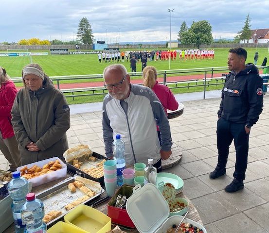 Mehrere Menschen stehen bei einem Tisch mit Essen. Ein älterer Mann mit Brille schaut in Richtung des Feldes, wo sich eine Fußballmannschaft aufwärmt.