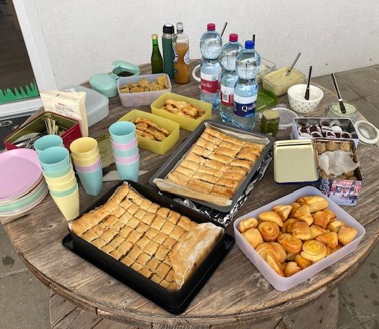 Ein Holztisch mit einer Vielzahl von Lebensmitteln wie Gebäck, Getränken und Gewürzen. Schüsseln, Teller und Tassen sind herum verstreut.
