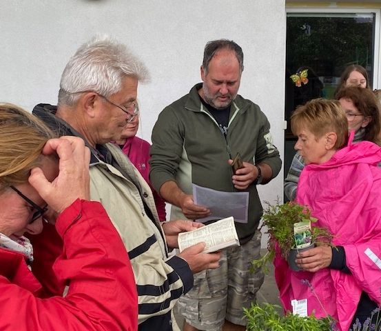 Eine Gruppe von Erwachsenen in Jacken, einige mit Pflanzen, versammelt sich draußen in der Nähe einer weißen Wand mit einem Schmetterling und einer Glastür im Hintergrund.