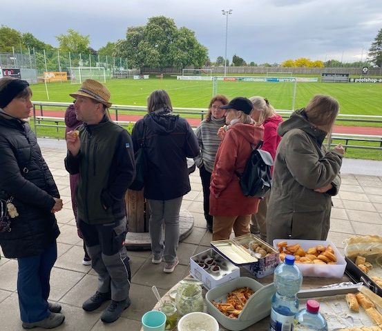 Eine Gruppe von Menschen versammelt sich bei einem Outdoor-Event in der Nähe eines Sportfelds, isst und unterhält sich. Tabletts mit Essen und Getränken stehen auf einem Tisch.