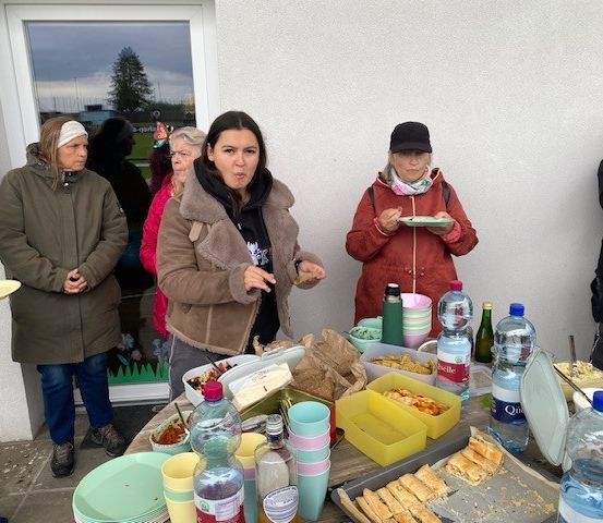 Eine Gruppe von Menschen versammelt sich um einen Tisch mit Essen und Getränken. Eine Frau in einer braunen Jacke steht mit einem Teller. Hinter ihr hält eine weitere Frau einen Teller. In der Nähe isst eine Frau in einer roten Jacke. Flaschen und Becher stehen auf dem Tisch. Ein Gebäude mit einem Glasfenster ist im Hintergrund.