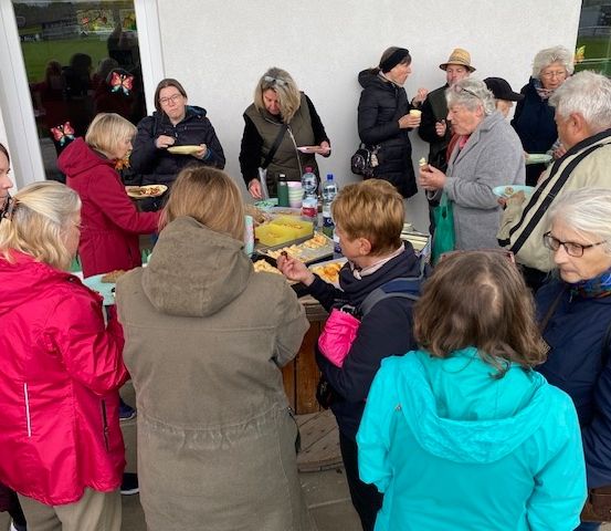 Eine Gruppe von Menschen versammelt sich um einen Tisch und isst und plaudert vor einem Gebäude. Sie tragen Wintermäntel und einige haben Hüte. Eine Frau in einem roten Mantel hält einen Teller.