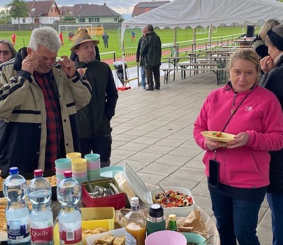 Eine Gruppe von Menschen steht um einen Tisch mit Essen und Getränken. Ein älterer Mann in einer beigen Jacke isst etwas. Eine Frau in einer rosa Jacke hält einen Teller. Ein Mann in einem Hut steht in der Nähe. Im Hintergrund ein Rasenfeld und Häuser.